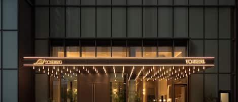 Ein moderner Hotelaufgang mit Glasfassade, beleuchteter Beschilderung und Pflanzen.