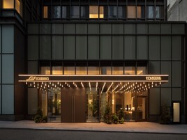 A modern hotel entrance with a glass facade, illuminated signage, and outdoor plants.