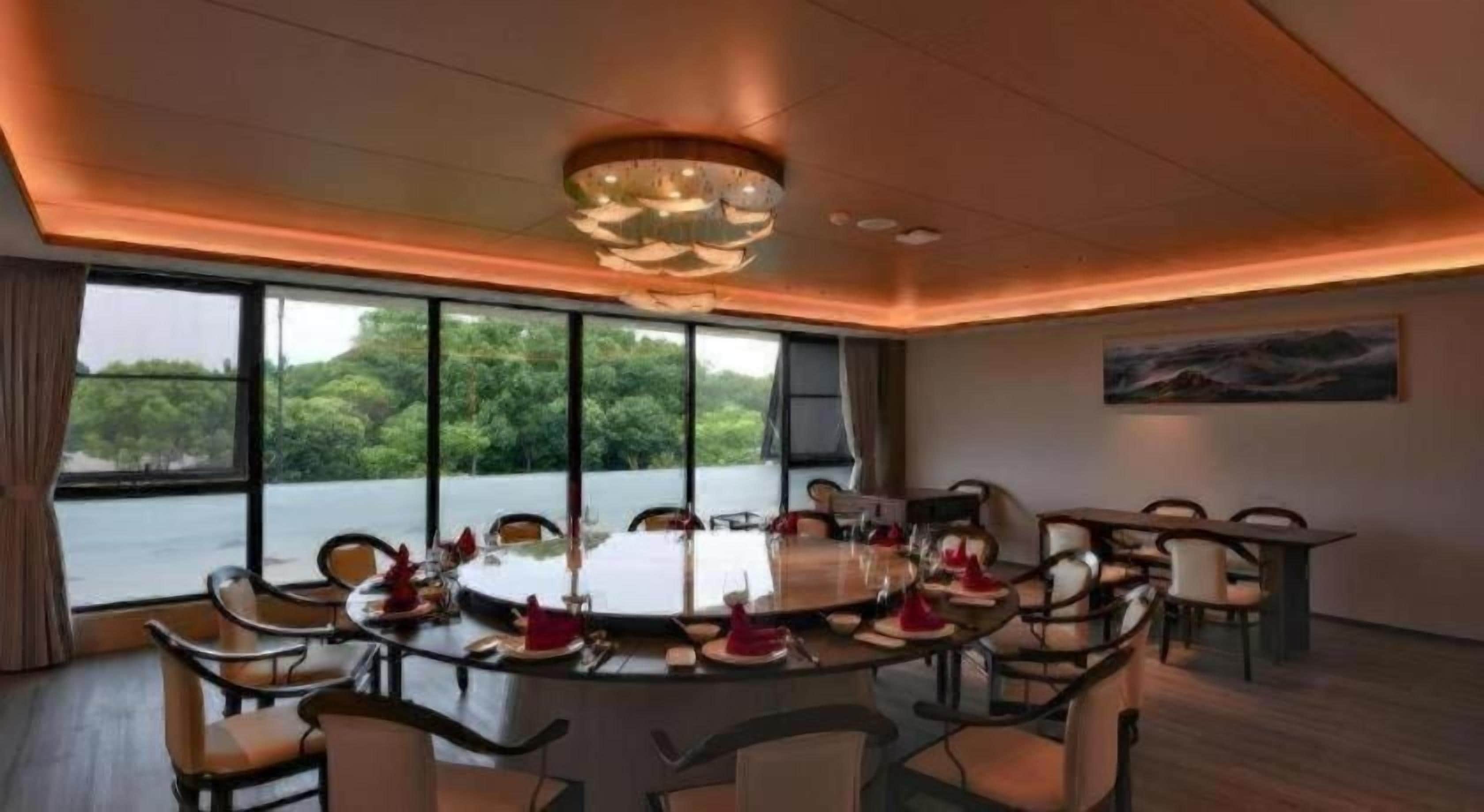 A dining area with a round table set for multiple guests, large windows offering a view of greenery, and a modern chandelier.