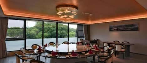 A dining area with a round table set for multiple guests, large windows offering a view of greenery, and a modern chandelier.