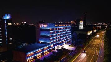 Un hotel de varios pisos con señalización iluminada, un estacionamiento y un paisaje urbano al fondo.