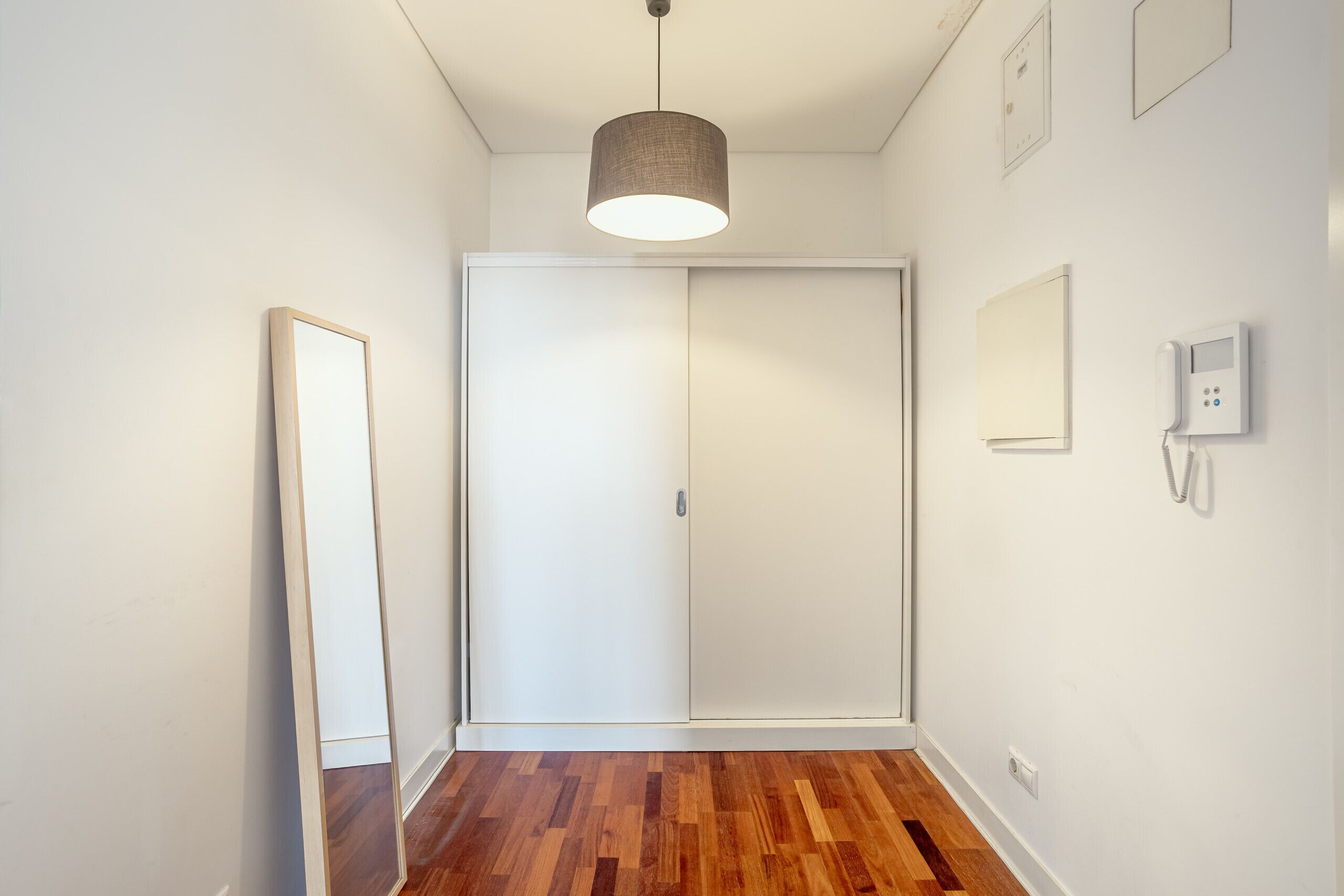 A hallway with a wooden floor, a white door, a hanging light fixture, and a wall-mounted phone.