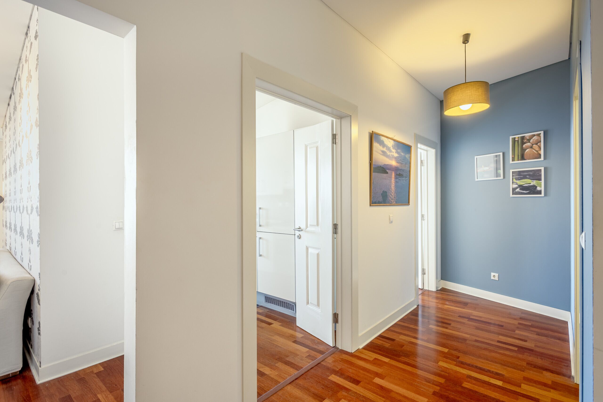 A hallway with wooden flooring, a white door, and a blue wall with framed pictures.