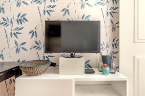 Ein Flachbildfernseher auf einem weißen Standfuß, ein gewebter Korb und dekorative Vasen auf einem weißen Schreibtisch vor einer Wand mit blauem Blattmuster.