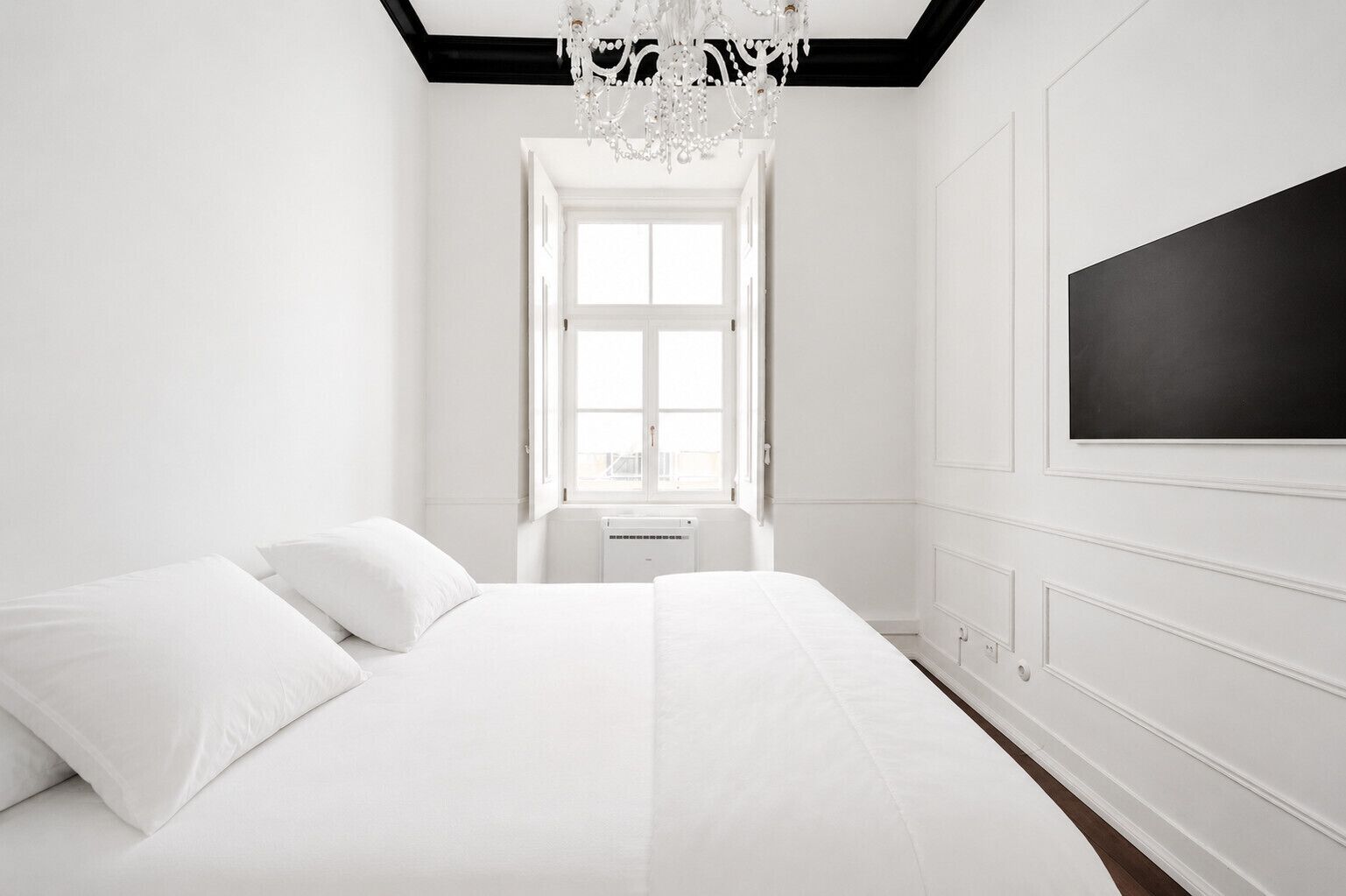 A white bedroom with a large bed, a chandelier, a window, and a wall-mounted TV.