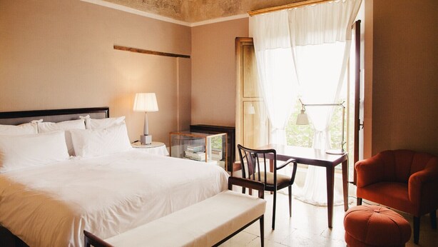 Una habitación de hotel con cama, mesita de noche, lámpara, silla y ventana con cortinas.