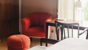 Habitación de hotel con un armario de madera, un sillón rojo, una mesa de madera con una lámpara y una cama blanca.