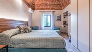 Quarto com teto de tijolos, cama com almofadas estampadas, janela com vista e um pequeno espaço para sentar.