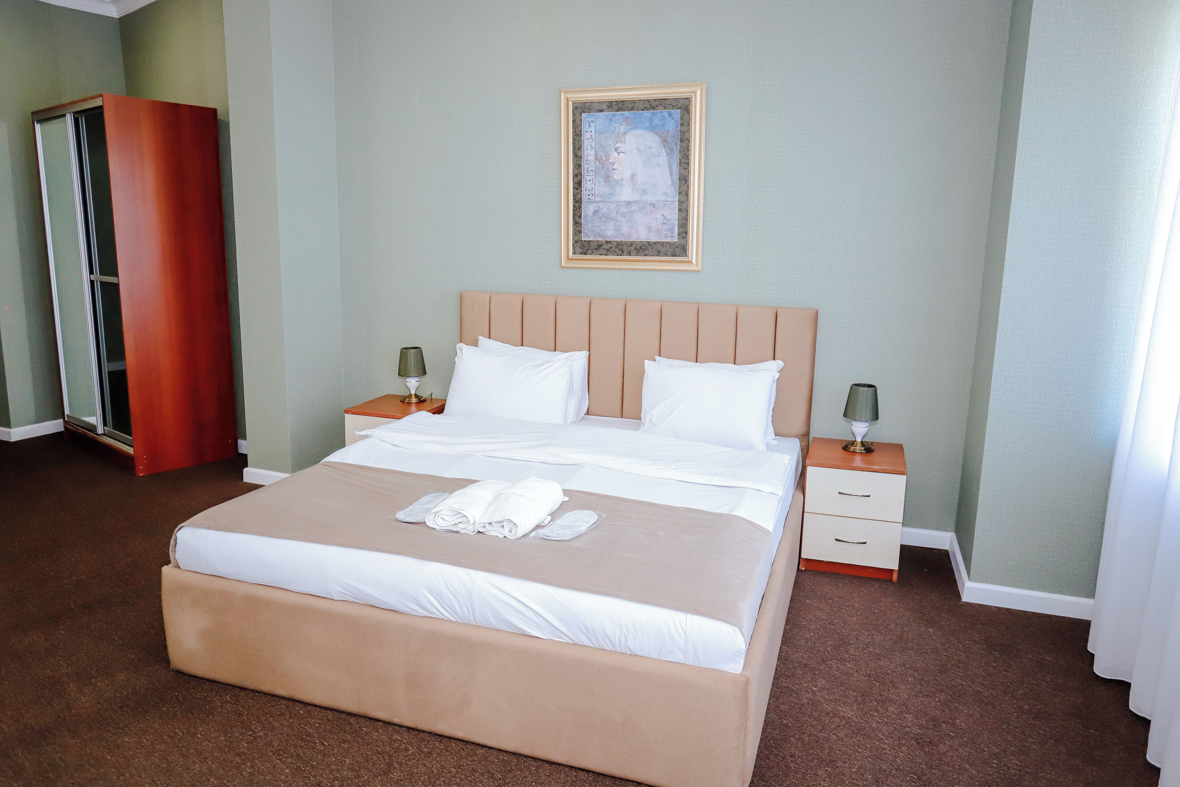 A hotel room with a large bed, two bedside tables, a wardrobe, and a painting on the wall.