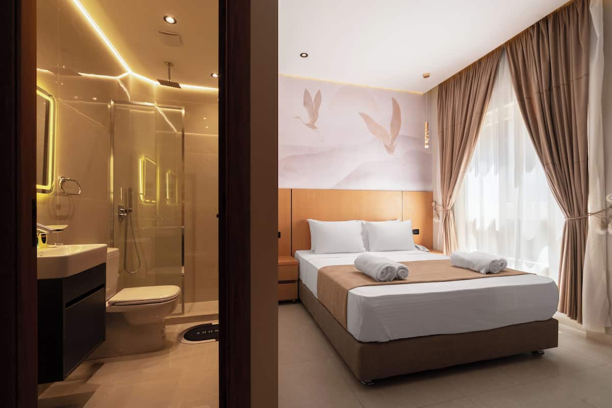 A modern hotel room with a glass-enclosed shower, a separate toilet, a large bed with white linens, and a large window with curtains.