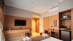 Ein modernes Hotelzimmer mit einem Bett, einem an der Wand befestigten Fernseher, einem Schreibtisch mit Stuhl, einem kleinen Kühlschrank und einem durchgehenden Türspalt einsehbarer Badezimmer.