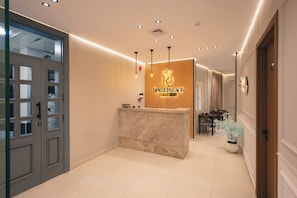Zona de recepción de hotel con un mostrador de mármol, un cartel que dice 'Royal Palace Hotel' y un comedor con mesas y sillas.