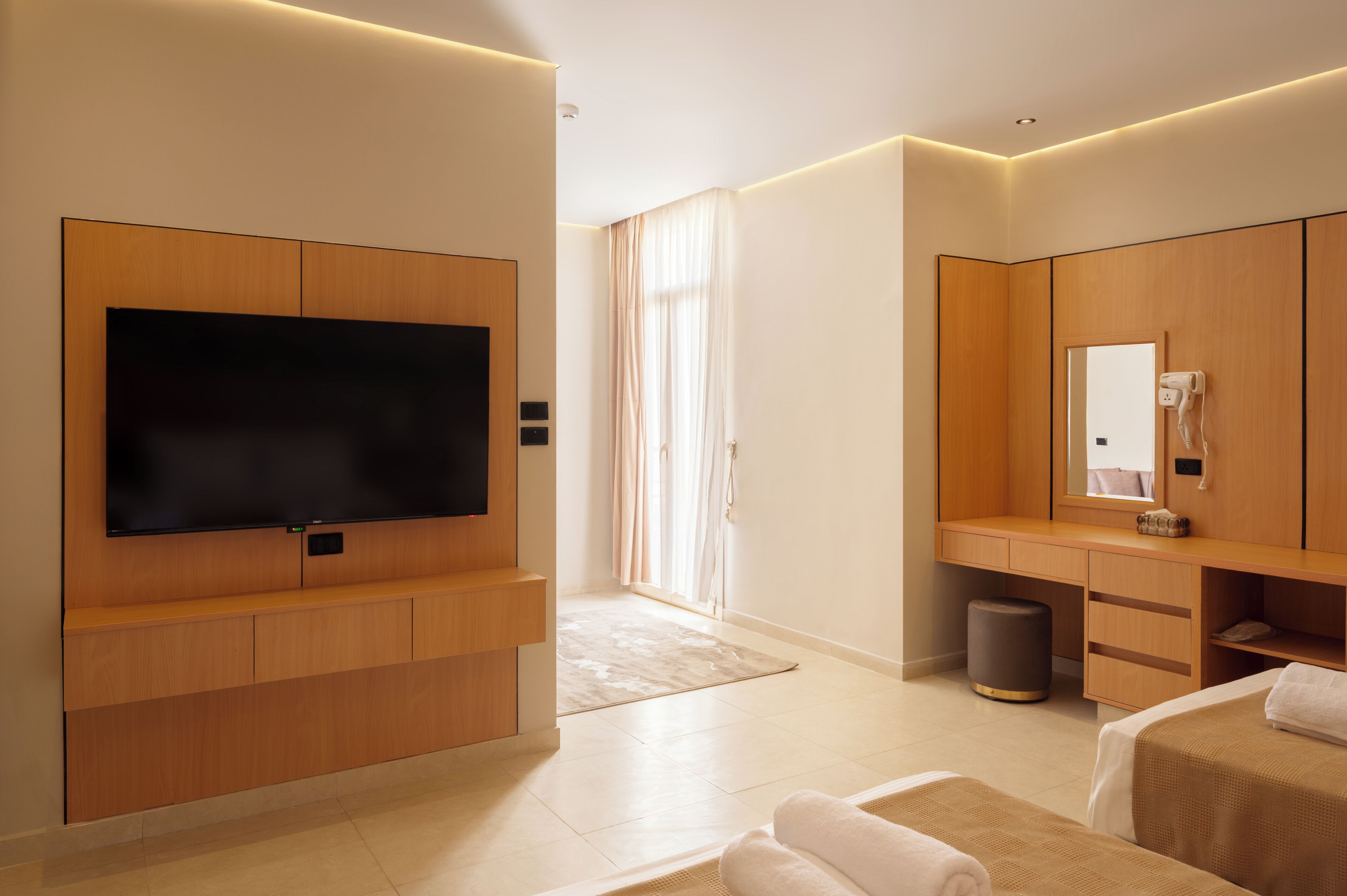 A modern hotel room with a flat-screen TV, wooden furniture, a large window with curtains, and a trash bin.