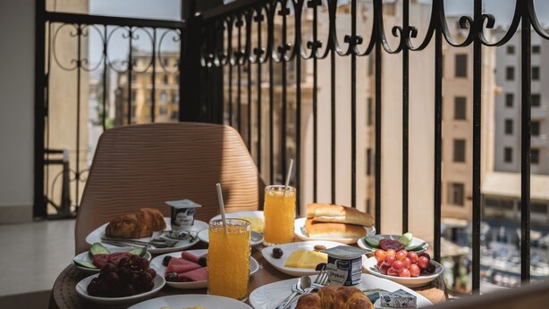 Ein Balkon mit Frühstücksbuffet, darunter Croissants, Eier und Saft, mit Blick auf eine Stadtlandschaft.