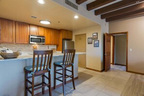 A kitchen with wooden cabinets, a microwave, a refrigerator, and two bar stools.