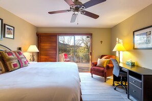 A bedroom with a bed, a desk, a chair, a ceiling fan, and a balcony with a view.