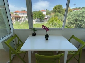 Ein Balkon mit einem weißen Tisch, zwei Stühlen, zwei Kübelpflanzen und Blick auf Häuser und Grünflächen.