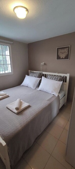 A bedroom with a bed, pillows, a bedside table, a wall-mounted light, and a window. - Villa- Nea Kalikrateia - 2-bdroom Apartment (Nea Propontida)