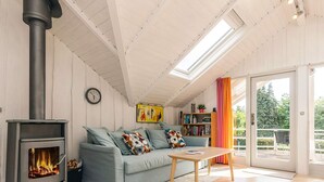 Una sala de estar acogedora con una estufa de leña, un sofá con cojines estampados, una estantería y un ventanal grande.