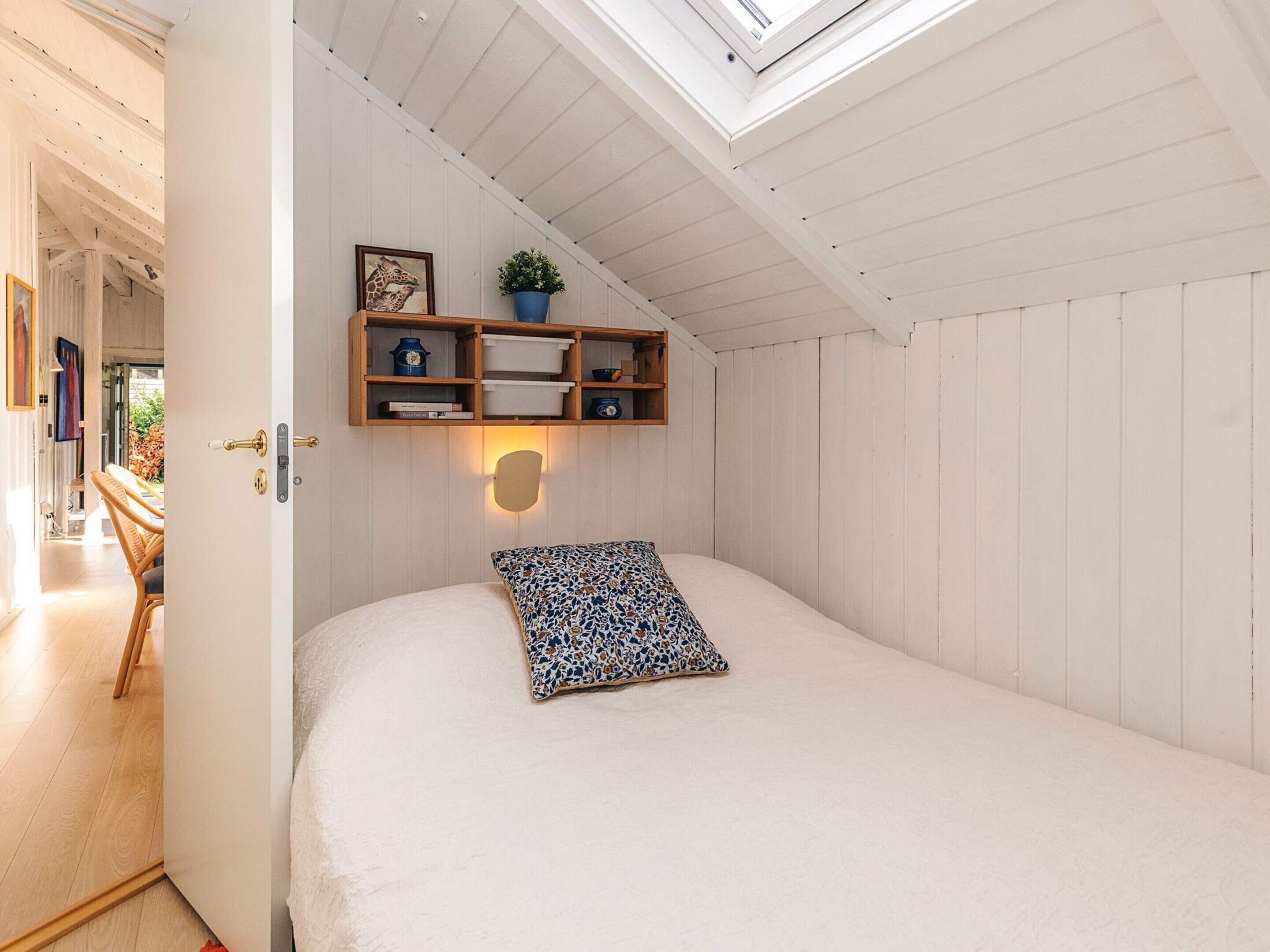 Une chambre avec un puits de lumière, un lit agrémenté d’un coussin à motifs, une étagère en bois avec des livres et une plante, et un luminaire fixé au mur.