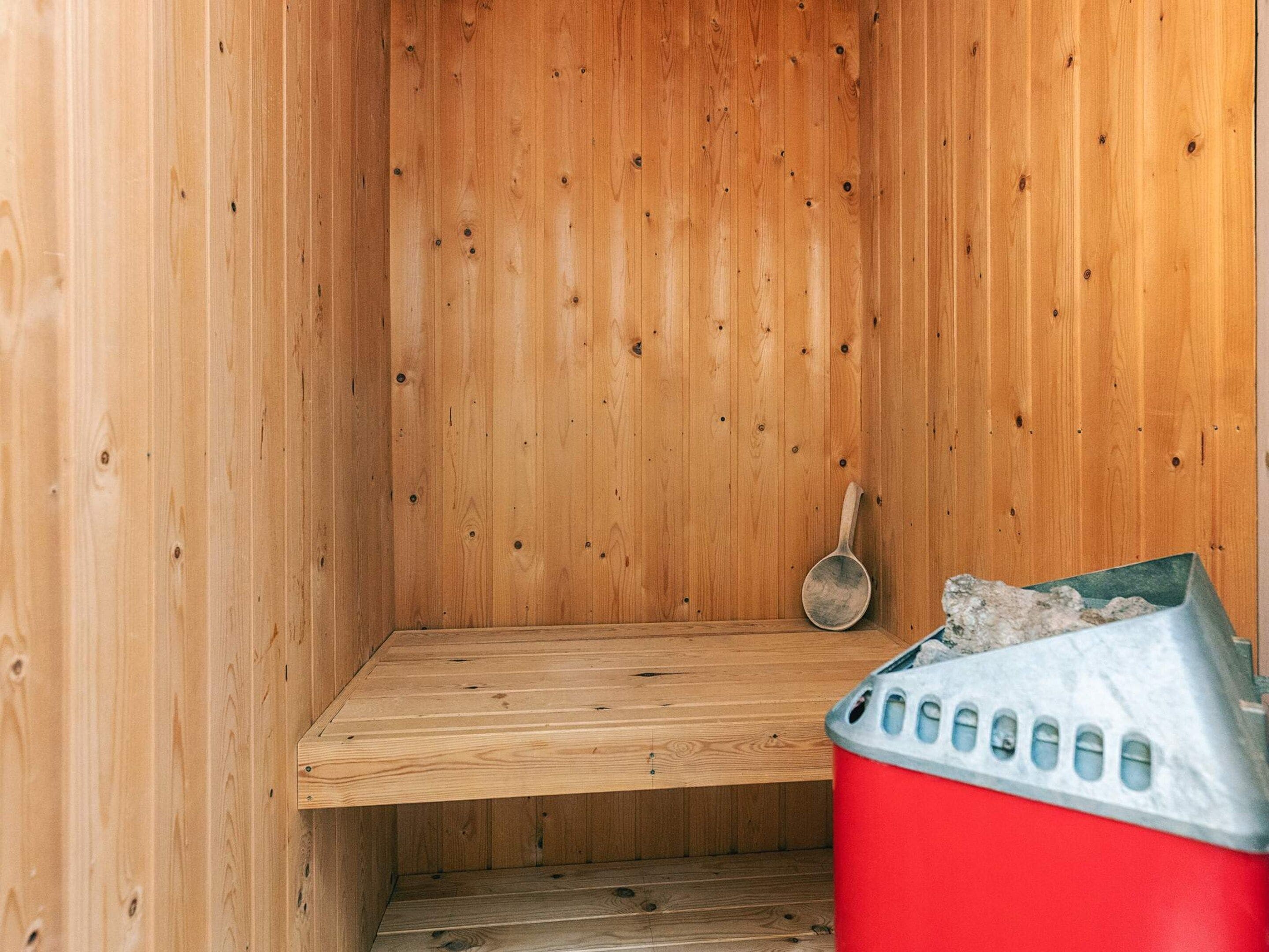 A sauna with a wooden interior, a bench, a metal ladle, and a red trash bin.