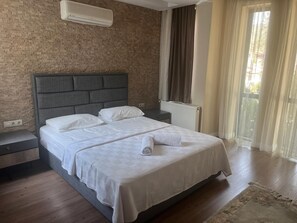 A bedroom with a brick wall, a grey headboard, a white bed with a checkered duvet, a wooden nightstand, and a window with curtains.
