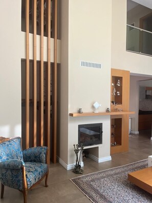 A living room with a blue patterned armchair, a fireplace, a wooden shelf with decorative items, and a large window.