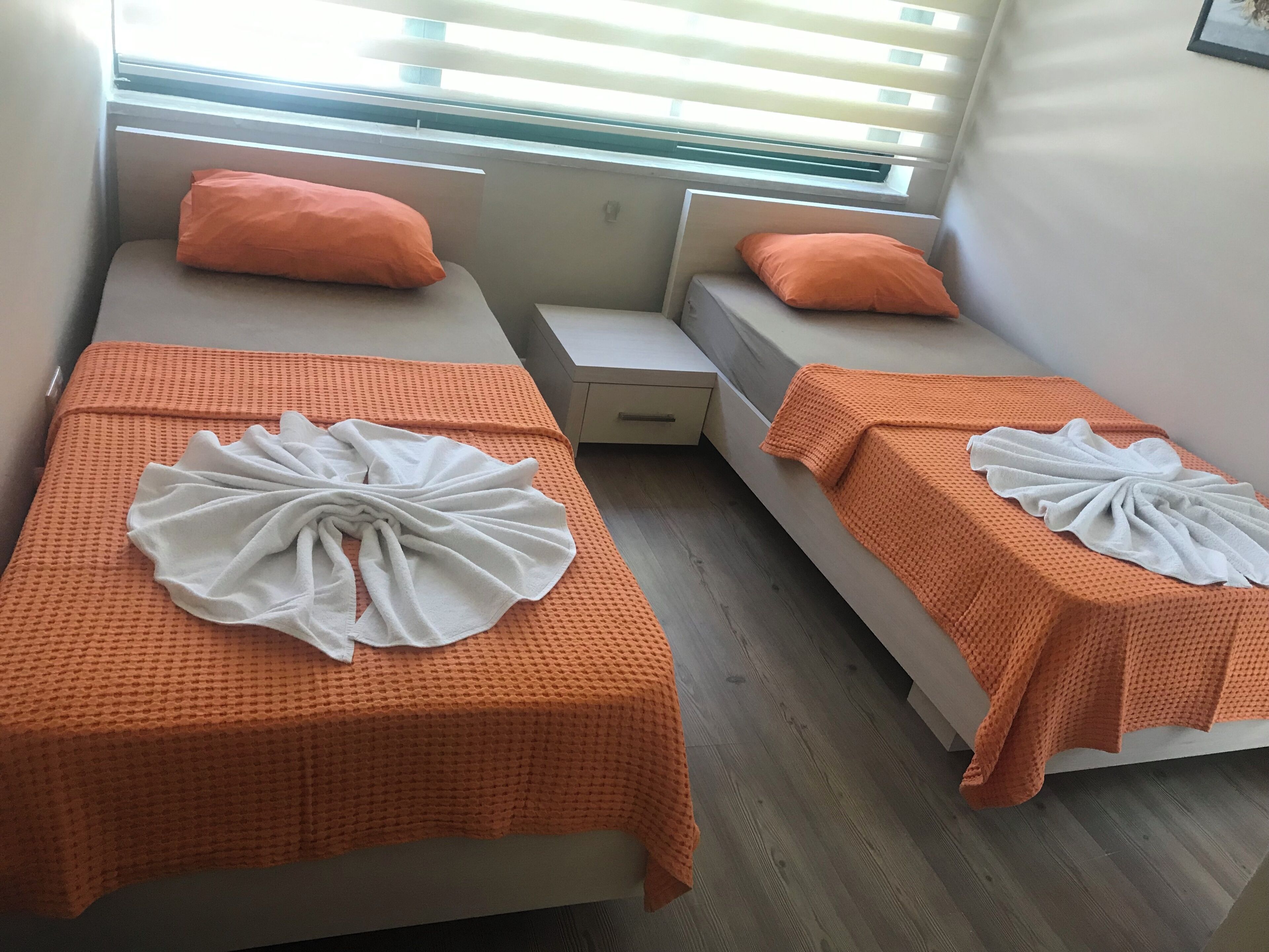 Twin beds with orange blankets and white towels, a small bedside table, and a window with blinds.