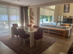 A modern living room with a glass dining table, red chairs, a sofa, a TV, and a large window with a view of greenery.