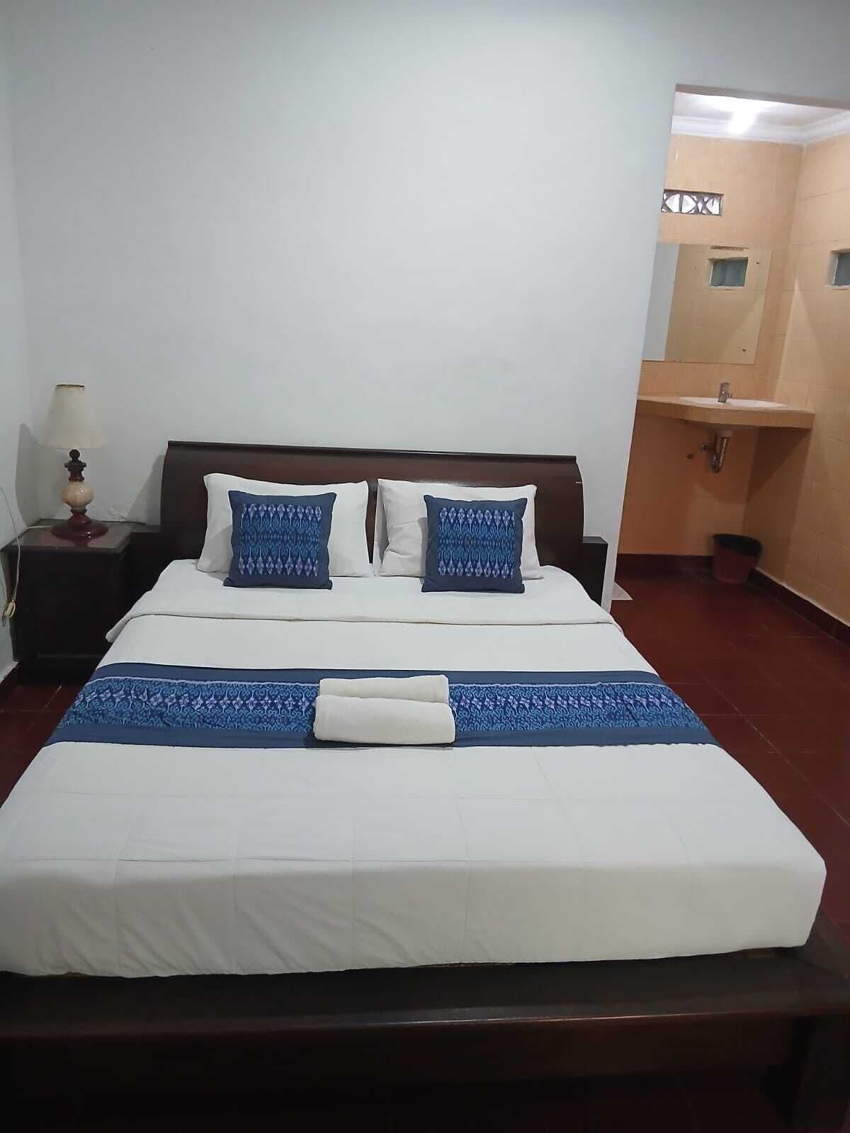 Habitación de hotel con cama, mesita de noche, lámpara y baño con lavamanos.