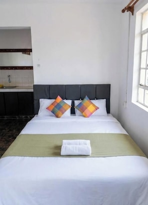 A neatly made bed with white linens and two colorful pillows, a green throw blanket, and a window with a view of the outside.