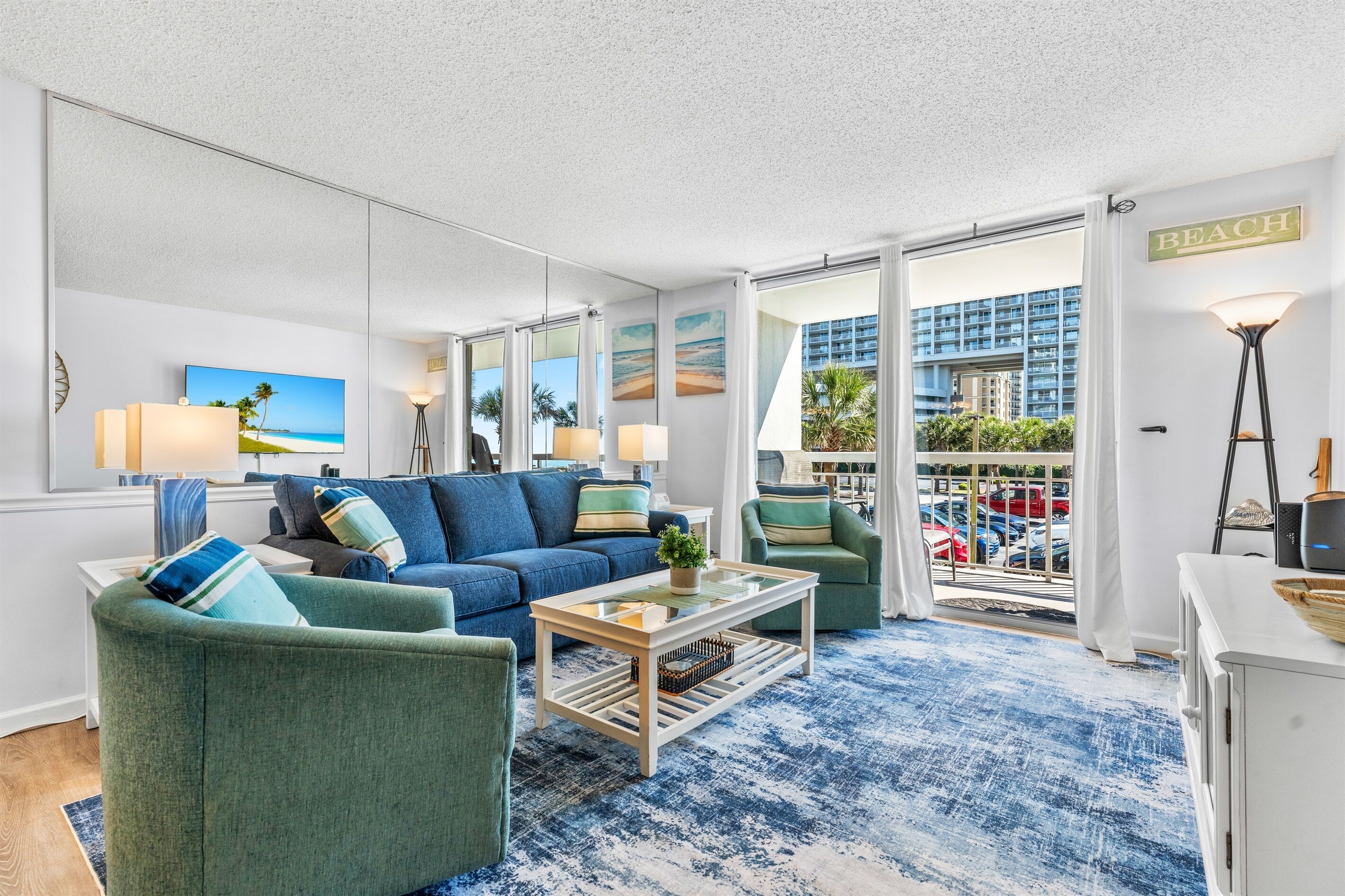 Un salón con una decoración en tonos azul y blanco, un ventanal con vistas a un edificio y coches estacionados, y un cartel que dice 'BEACH'.
