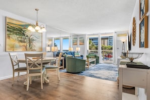 A spacious living area with a dining set, a green armchair, and a view of the beach through large windows. - South Hampton 107 Oceanview w/Pool & Beach (Myrtle Beach)