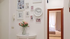Ein Flur mit weißer Wand, einer runden Uhr, einem kleinen Tisch mit Blumen in einer Vase und einer Tür, die in ein Schlafzimmer führt.