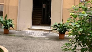 Ein Gebäudeeingang mit einer Tür, die mit einem 'M' gekennzeichnet ist, einer Treppe, die nach innen führt, und Pflanzgefäßen auf dem Bürgersteig.