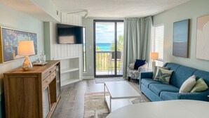 A living room with a sofa, a coffee table, a TV mounted on the wall, and a sliding glass door leading to a balcony with a view of the ocean.
