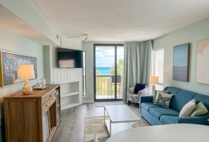 A living room with a sofa, a coffee table, a TV mounted on the wall, and a sliding glass door leading to a balcony with a view of the ocean.