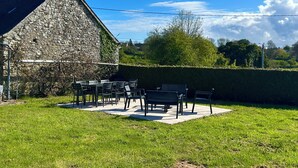 A stone building with a sloped roof, a grassy yard, and a patio with chairs and tables. - Les figuiers Charming stone house Pets welcome (Gonneville-Le Theil)
