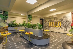 A lobby with a curved reception desk, yellow tables, green plants, and a wall with a 'HELLO' sign. - The Hosteller Bangalore, CV Raman Nagar (Bengaluru)