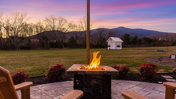 A stone fire pit with a wooden bench, a white building in the background, and a grassy field with trees and mountains. - Afton Awaits (Afton)