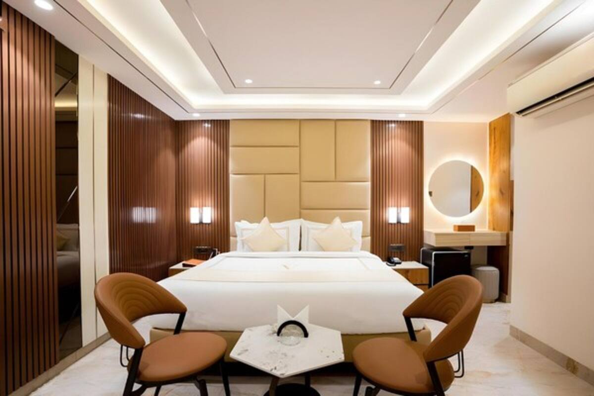 A modern hotel room with a large bed, two armchairs, a small table, a round mirror, and a wooden panel wall.