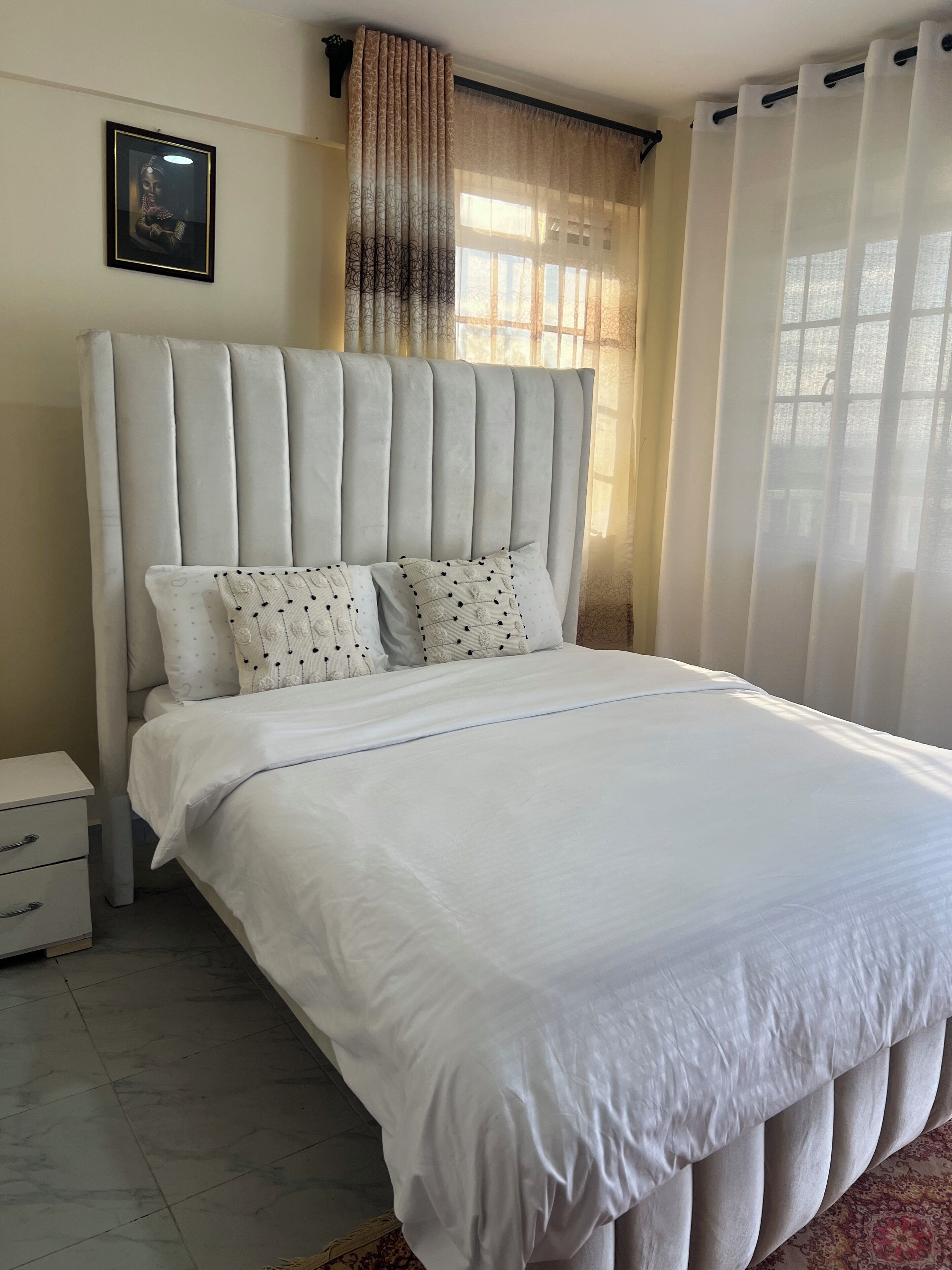 A bedroom with a large bed, a nightstand, a framed picture on the wall, and sheer curtains.
