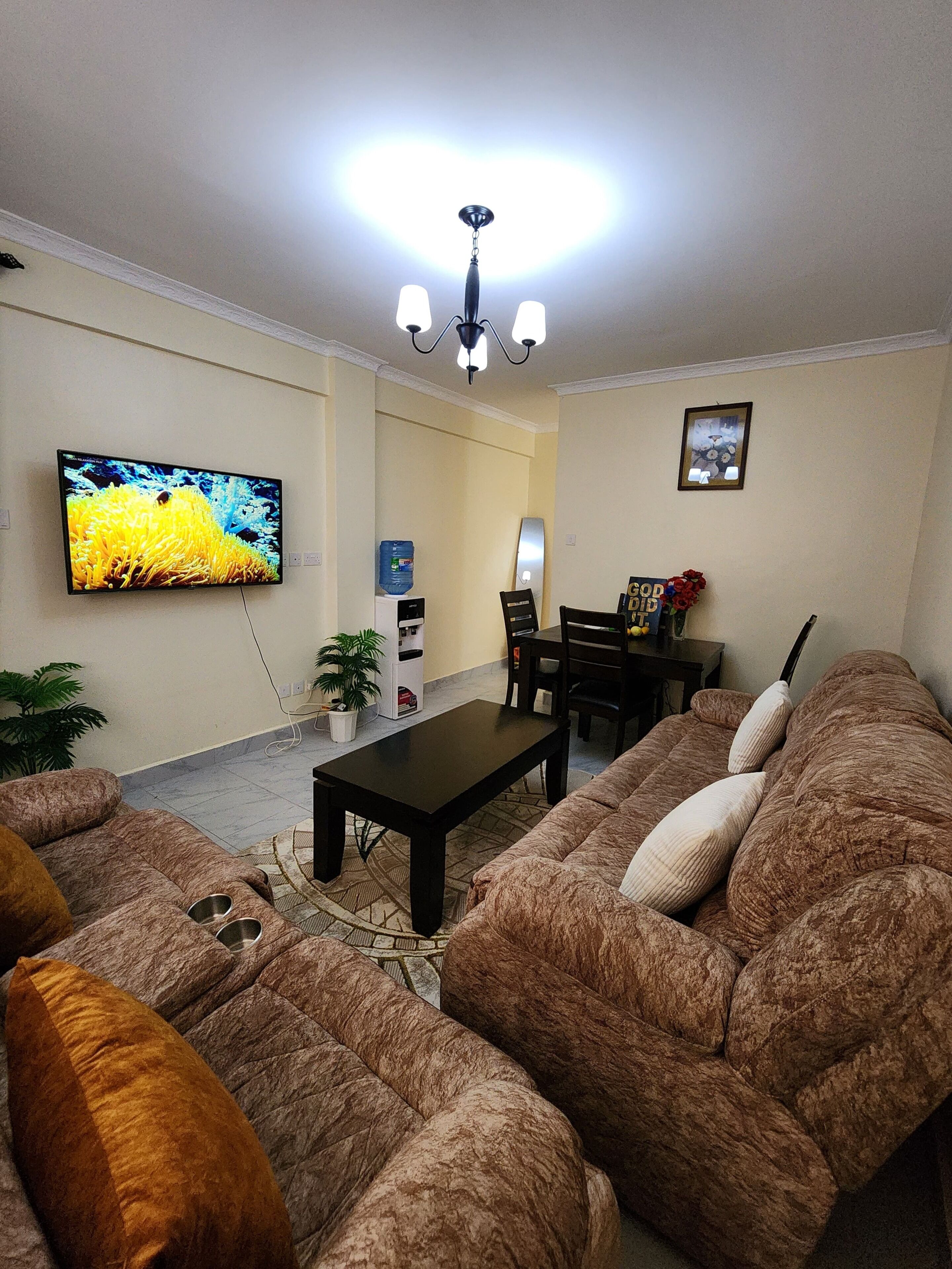 A living room with a flat-screen TV, a coffee table, a sofa, a dining table with chairs, and a water dispenser.
