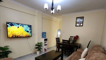 A living room with a flat-screen TV, a coffee table, a sofa, a dining table with chairs, and a water dispenser.