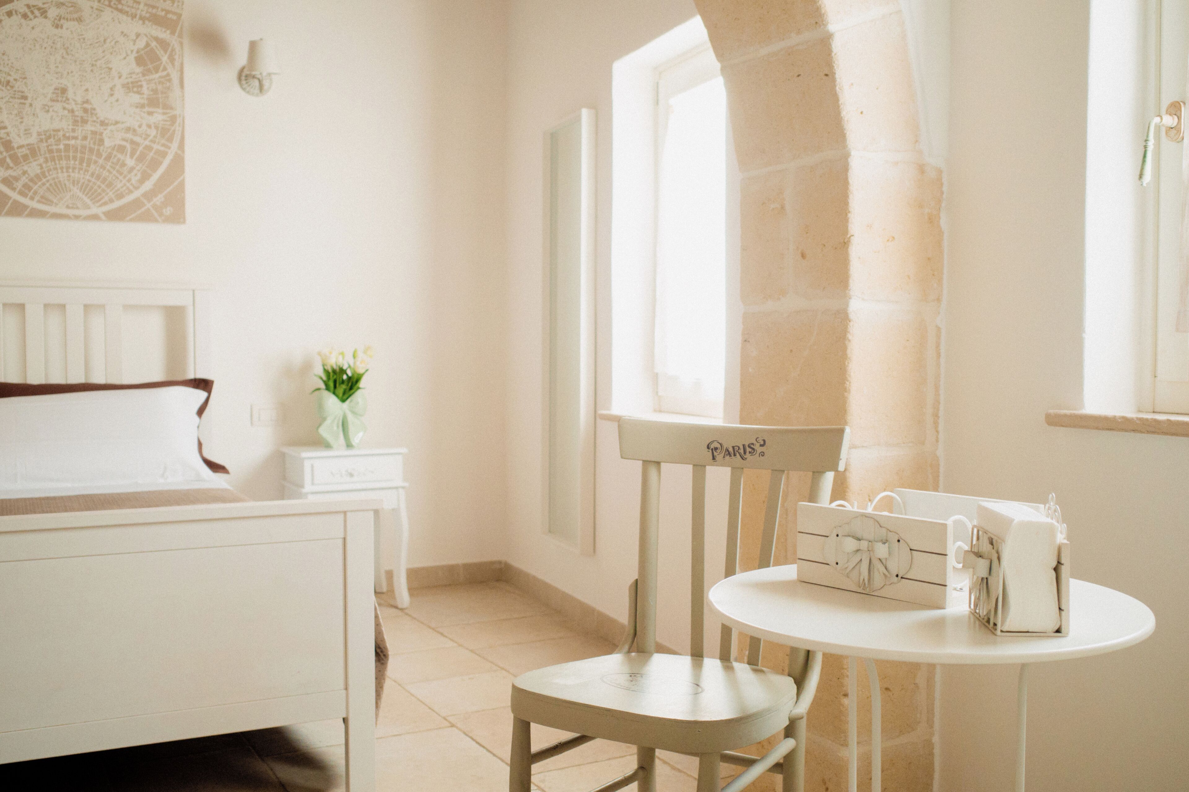 Una habitación con cama, una silla, una mesa con un adorno, un florero con flores y una ventana con vistas.