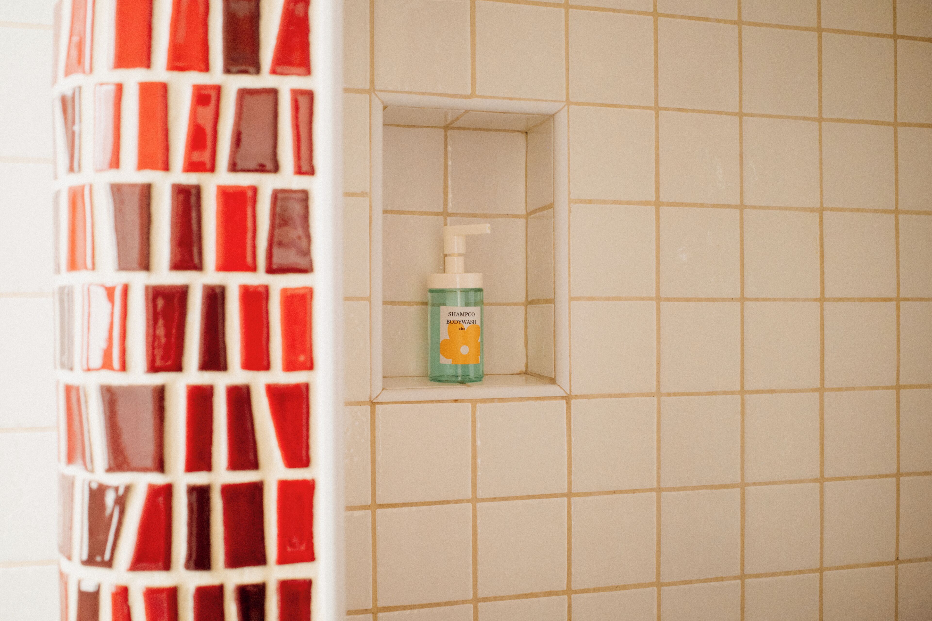 A bathroom with a tiled wall, a shower area, and a bottle of shampoo on a shelf.