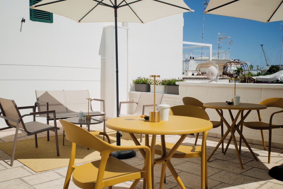 A terrace with yellow chairs and tables, a white wall, and a view of the sea and buildings in the background.