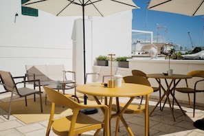 A terrace with yellow chairs and tables, a white wall, and a view of the sea and buildings in the background.