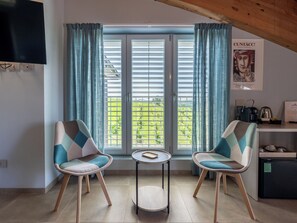 Una stanza con due sedie, un tavolino, una finestra con tende, una televisione e un quadro appeso al muro.