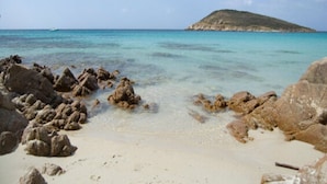 Een strand met helder turquoise water, rotsformaties en een heuvel in de verte.